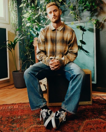 A fair-skinned man with red hair and a plaid shirt sits indoors surrounded by plants, a guitar, and music equipment.
