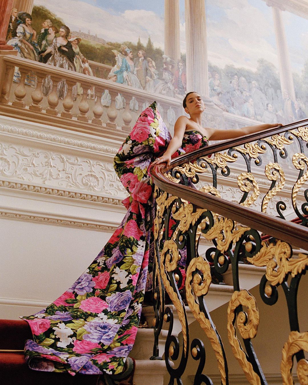 Woman poses on ornate staircase in floral gown, historic building. Red carpet. Mural with 18th-century figures. Gold and dark-brown metalwork.
