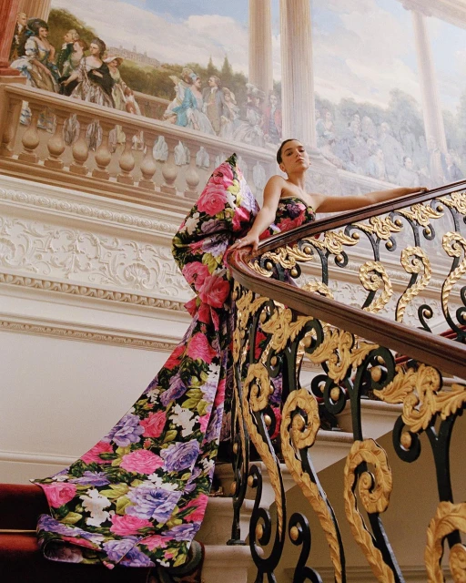 Woman poses on ornate staircase in floral gown, historic building. Red carpet. Mural with 18th-century figures. Gold and dark-brown metalwork.