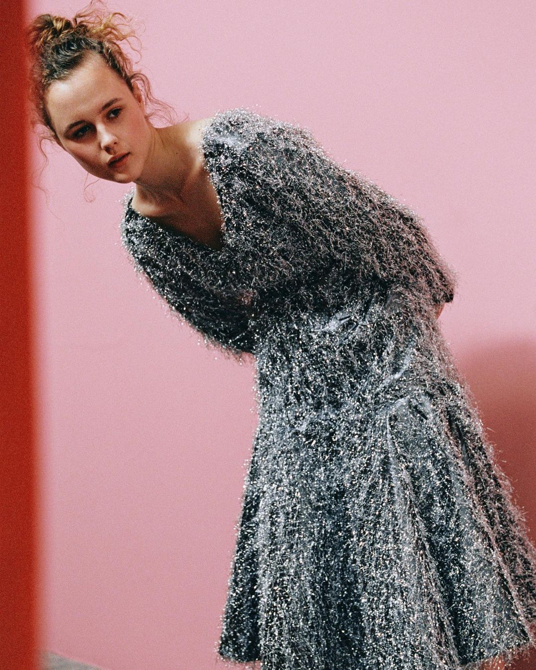 A young woman poses in a sparkly silver dress against a pink backdrop, creating a fashion-forward and visually striking composition.