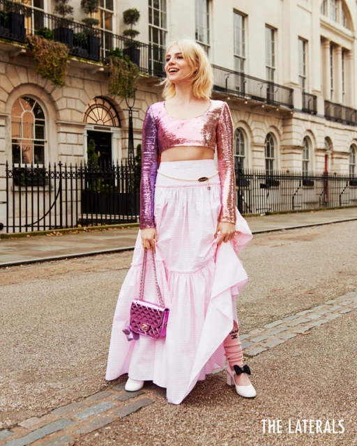 A full shot of a blonde adult female in a sequin crop top and light pink skirt standing on a city street with buildings in the background.