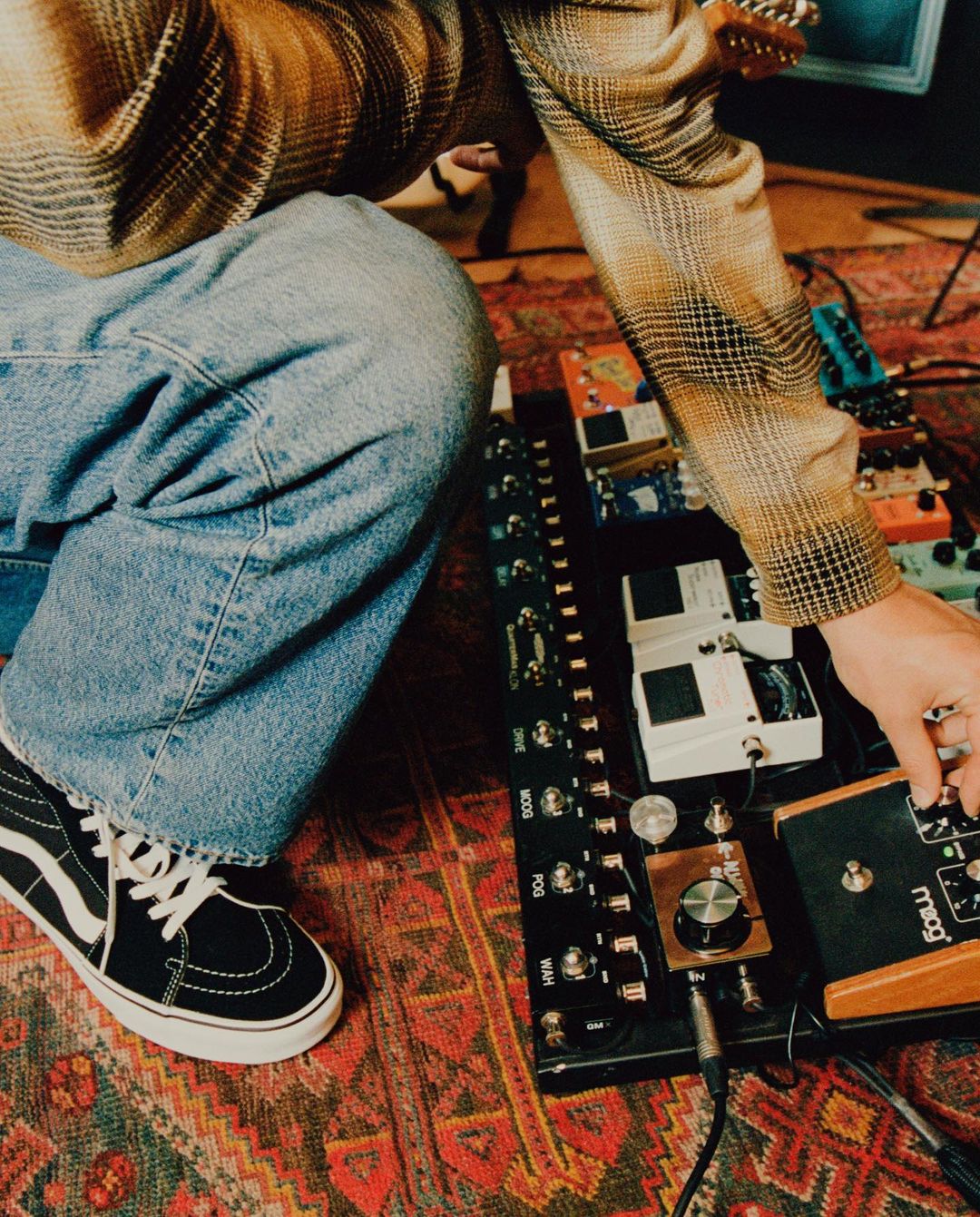 Unidentifiable musician adjusting knobs on guitar effects pedal board; black and white Vans shoe visible on a patterned rug.