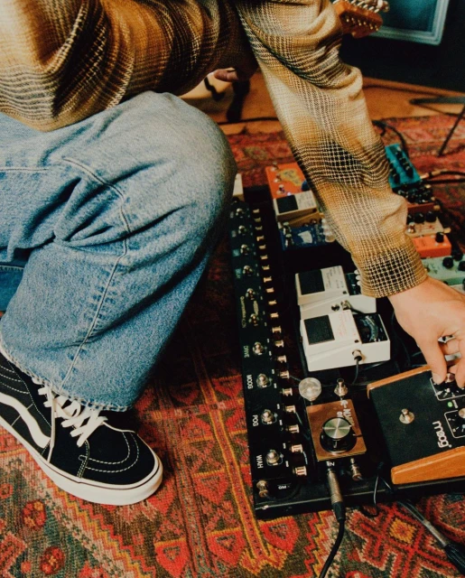 Unidentifiable musician adjusting knobs on guitar effects pedal board; black and white Vans shoe visible on a patterned rug.