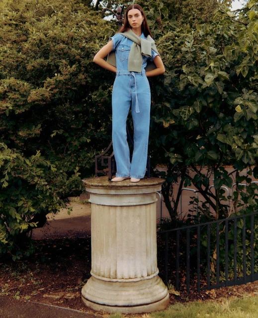 A woman stands on a stone plinth outdoors, wearing a denim jumpsuit and a sweater over her shoulders, in an eye-level shot.