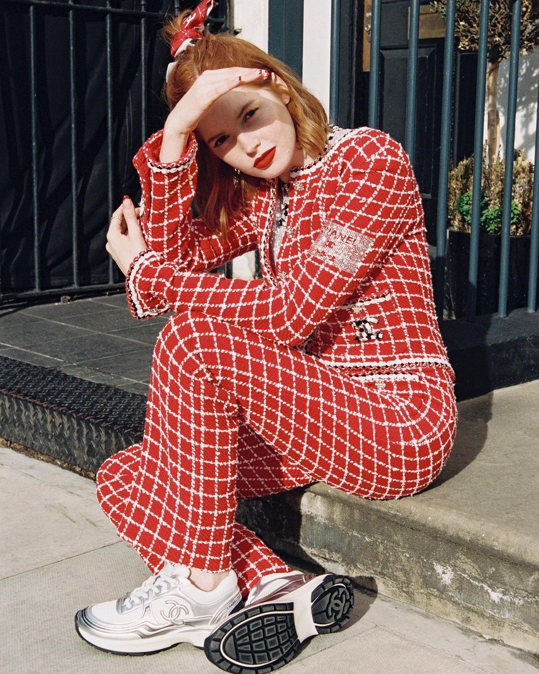 Adult female with red hair poses outdoors wearing a red and white checkered jacket and pants with sneakers while seated on a step.