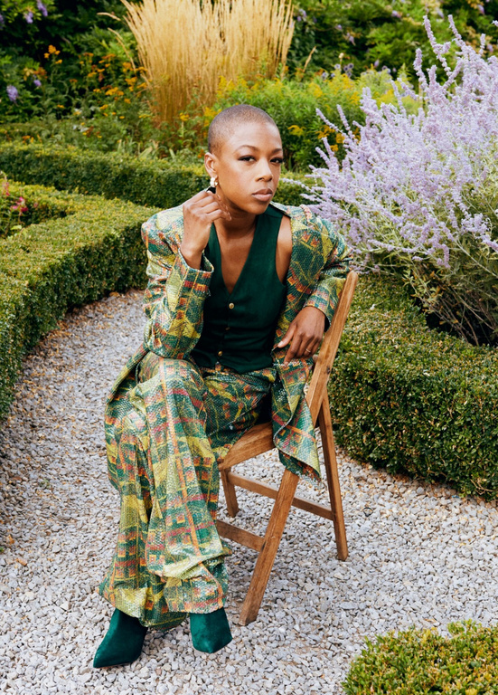 Stylish woman in a patterned suit sitting on a wooden chair in a garden setting with lush greenery and a gravel pathway.