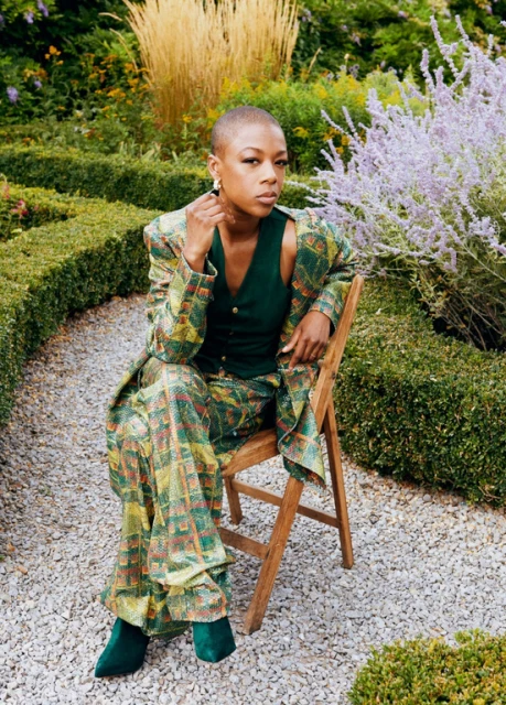 Stylish woman in a patterned suit sitting on a wooden chair in a garden setting with lush greenery and a gravel pathway.