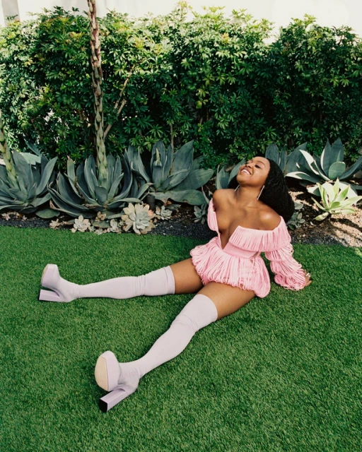 Woman in pink dress and thigh-high boots poses on grass with green foliage background, bathed in natural outdoor light.