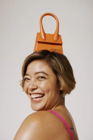 Woman with short brown hair smiling while balancing a small orange handbag on her head against white seamless backdrop