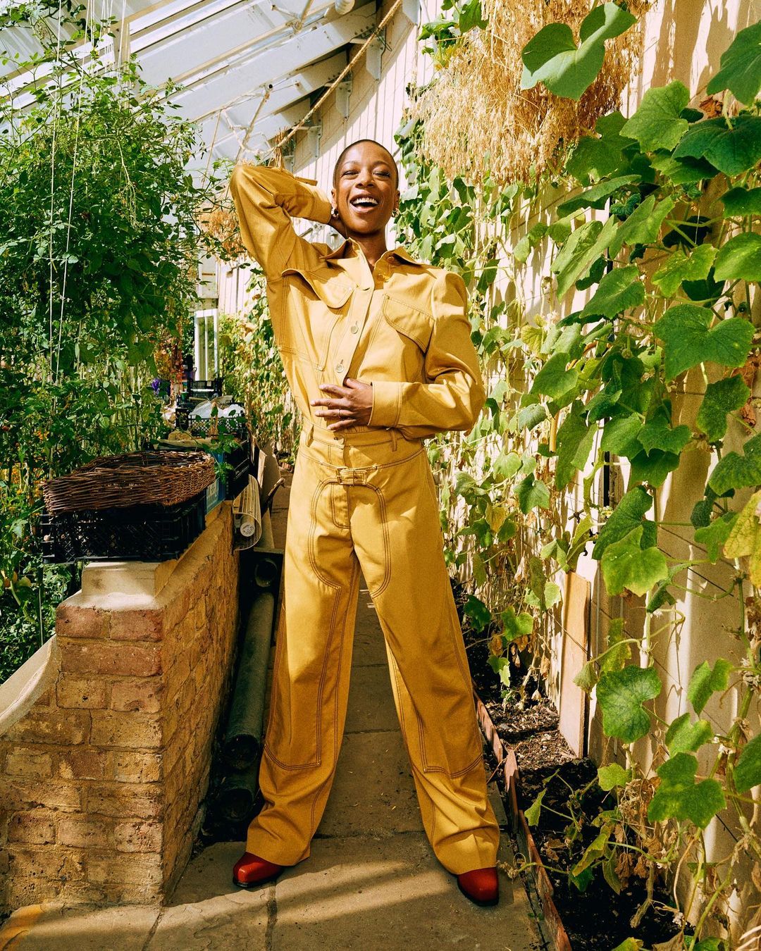 An adult woman in a gold-colored jumpsuit poses inside a greenhouse surrounded by vegetation, showcasing a blend of fashion and nature.