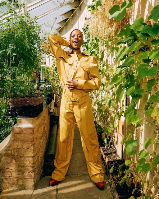 An adult woman in a gold-colored jumpsuit poses inside a greenhouse surrounded by vegetation, showcasing a blend of fashion and nature.