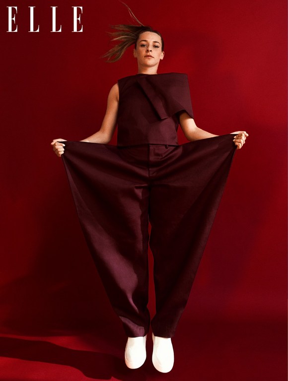 Fashion editorial shot of a woman in a maroon jumpsuit and white shoes jumping in front of a red backdrop with ELLE magazine logo.
