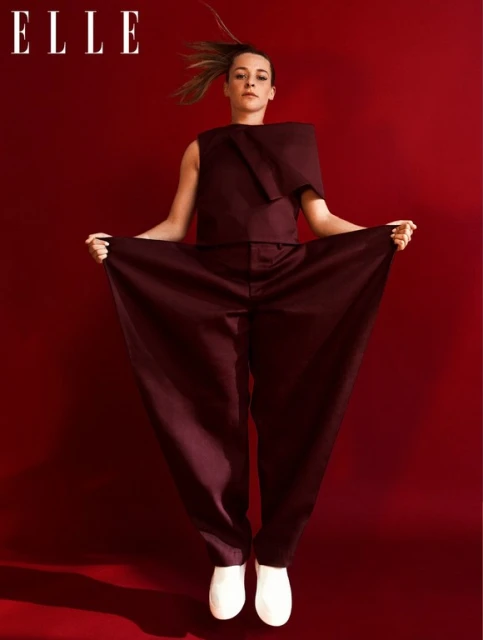 Fashion editorial shot of a woman in a maroon jumpsuit and white shoes jumping in front of a red backdrop with ELLE magazine logo.