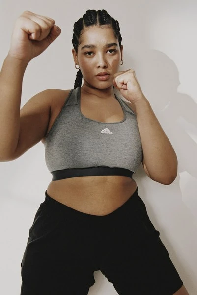 A woman in workout clothes stands in a fighting pose with a neutral expression, wearing a sports bra, shorts, and braided hair.