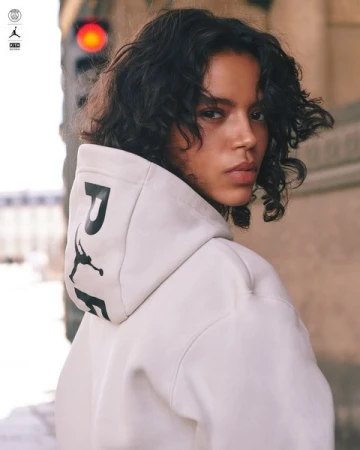 Close-up portrait of a woman with dark curly hair wearing an off-white hoodie, looking back over her shoulder outdoors.