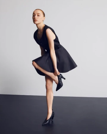 Woman in a black dress touching her heeled shoe against a neutral studio background