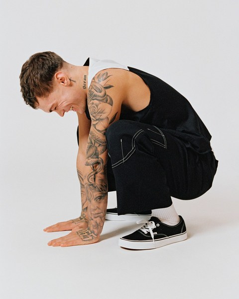 An adult male with visible tattoos poses in a crouched position, wearing a black vest, black pants, and black sneakers against a white background.
