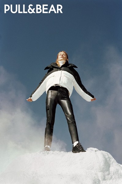 Model wearing black and white puffer jacket and black leather pants standing on white ground against clear blue sky.