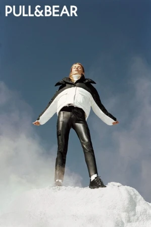 Model wearing black and white puffer jacket and black leather pants standing on white ground against clear blue sky.