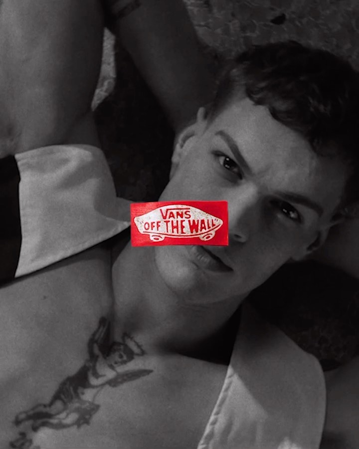 A shirtless, fair-skinned man is lying down in a photo studio, the Vans logo covering his eyes, and an angel tattoo on his chest.