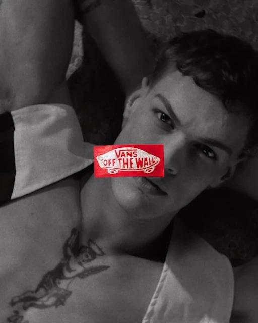 A shirtless, fair-skinned man is lying down in a photo studio, the Vans logo covering his eyes, and an angel tattoo on his chest.