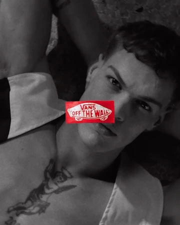 A shirtless, fair-skinned man is lying down in a photo studio, the Vans logo covering his eyes, and an angel tattoo on his chest.