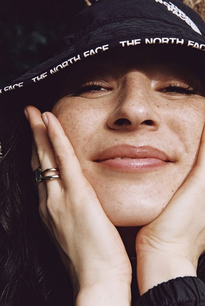 Close-up of a smiling woman with freckles wearing a black bucket hat with “The North Face” printed on it, holding her face with her hands.