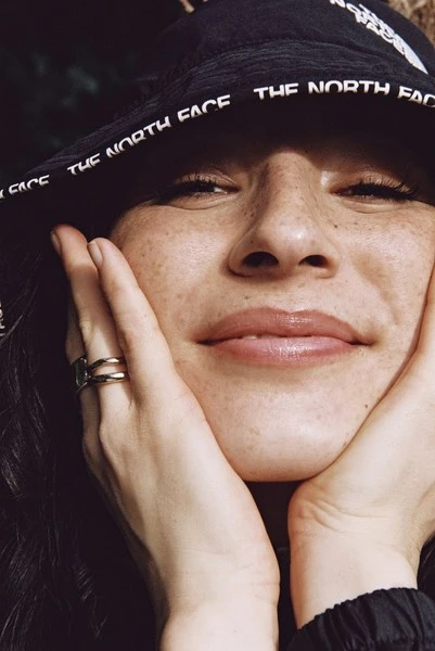 Close-up of a smiling woman with freckles wearing a black bucket hat with “The North Face” printed on it, holding her face with her hands.