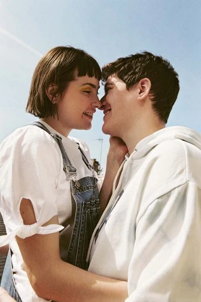 Close up of a happy couple outdoors with blue sky, wearing casual white tops and overalls, standing with noses touching.