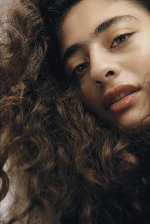 Close-up portrait of a woman with long dark curly hair and prominent eyebrows looking directly at the camera.