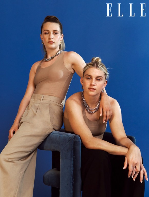Two adult women pose in a studio setting against a solid blue backdrop, wearing tan tops and chunky silver chain necklaces for a portrait.