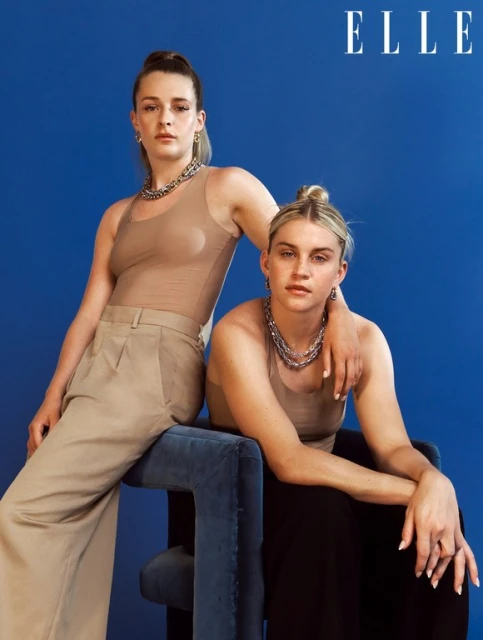Two adult women pose in a studio setting against a solid blue backdrop, wearing tan tops and chunky silver chain necklaces for a portrait.