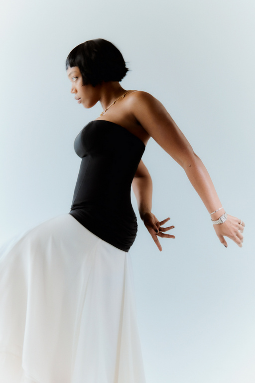 Elegant Black woman in black top and white skirt poses in a bright photo studio looking off camera with arms out.
