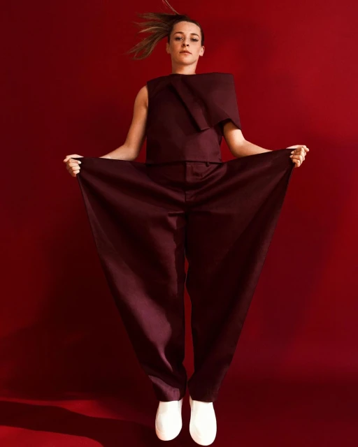 Woman in oversized auburn jumpsuit and white sneakers mid-jump against a scarlet backdrop in a studio setting for a fashion shoot.