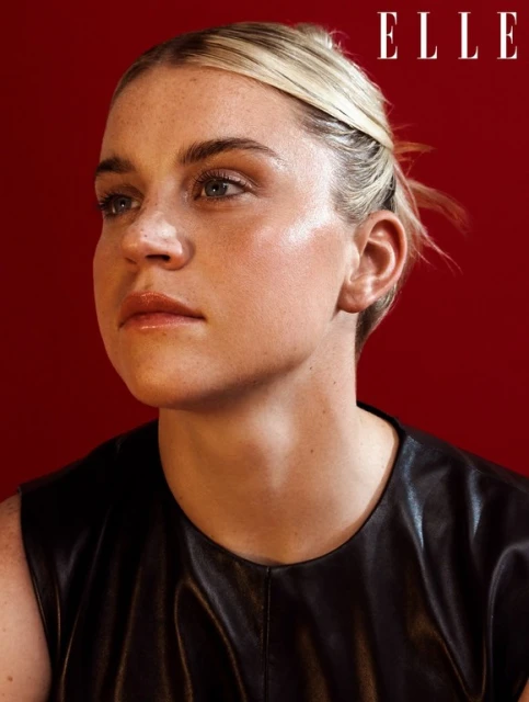 Close-up shot of a woman with blonde hair and wearing a black top, posing in front of a red background with the 'ELLE' logo visible.