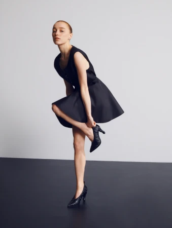 Model in a black sleeveless dress putting on a black heeled shoe against a studio backdrop.