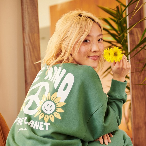 Blonde woman in a green sweatshirt holding yellow daisies, posing indoors, with a design of a peace sign and a sunflower on her back.