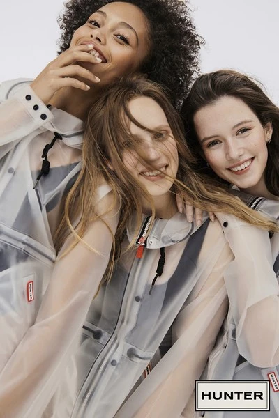 Three smiling young women wearing translucent Hunter raincoats against a white background, showcasing fashion and outdoor apparel.