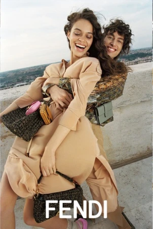 Two models embracing outdoors on a rooftop with city views, wearing beige attire and carrying patterned handbags, by photographer Rosaline Shahnavaz