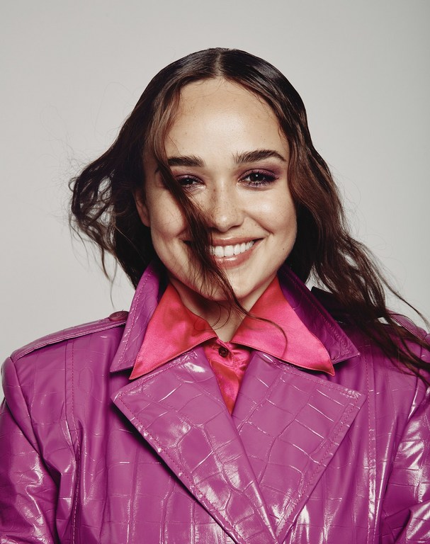 Close-up portrait of a smiling woman with brown hair, pink lipstick, and a magenta jacket against a neutral background.