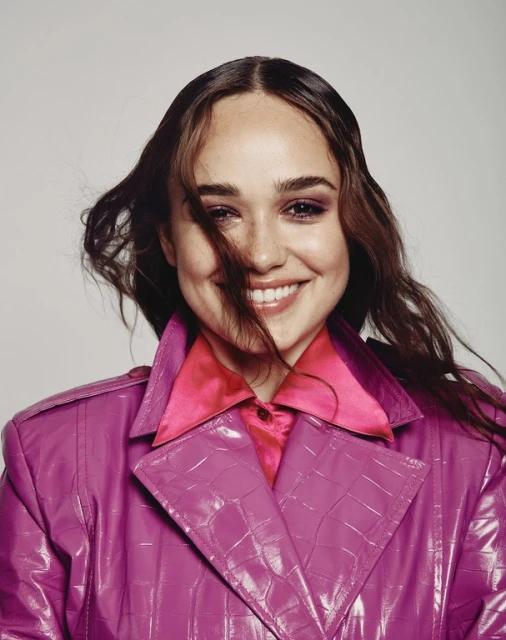 Close-up portrait of a smiling woman with brown hair, pink lipstick, and a magenta jacket against a neutral background.