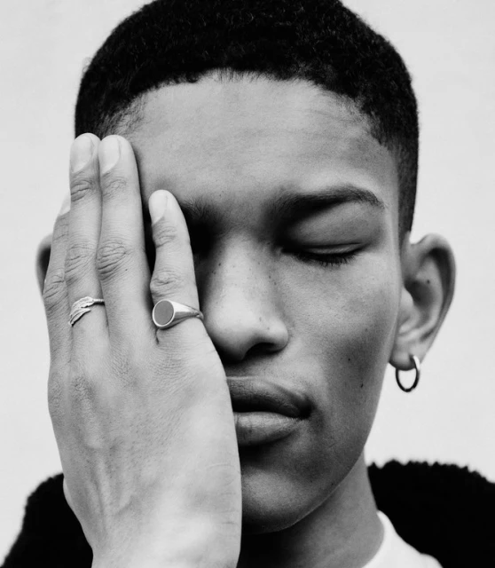 Close-up portrait of a young adult with closed eyes, featuring hand adorned with rings partially covering face; black and white.