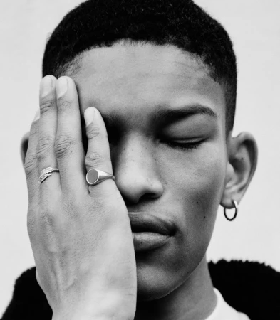 Close-up portrait of a young adult with closed eyes, featuring hand adorned with rings partially covering face; black and white.