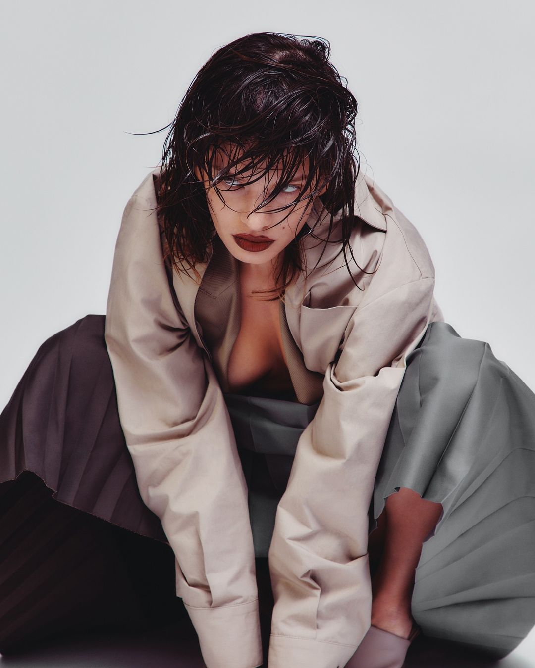 A woman with wet dark hair and dark lipstick poses in a tan coat and pleated skirt against a neutral background in a studio shot.
