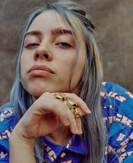 Close-up shot of a blue-haired adult female with gold rings on her hand, wearing a patterned blue shirt, against a grey wall. 