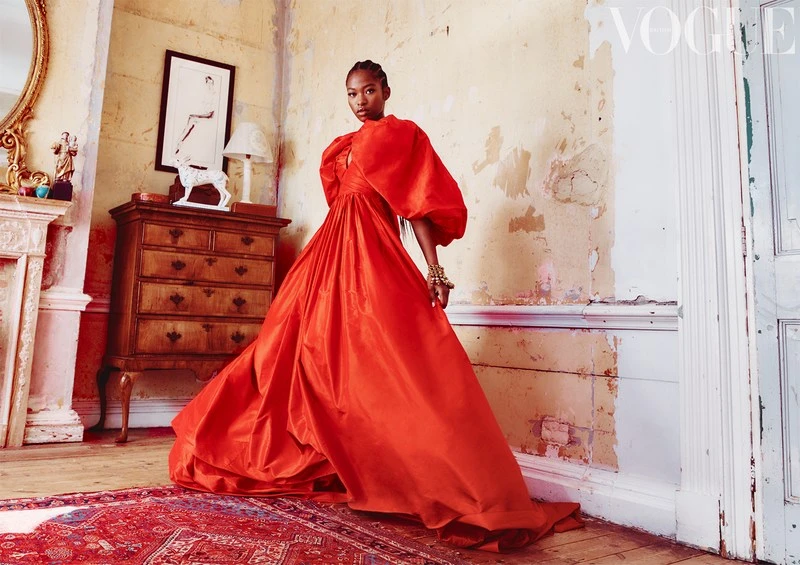 Fashion editorial shot of an African American woman in a voluminous orange gown, posing in an old room with vintage decor.