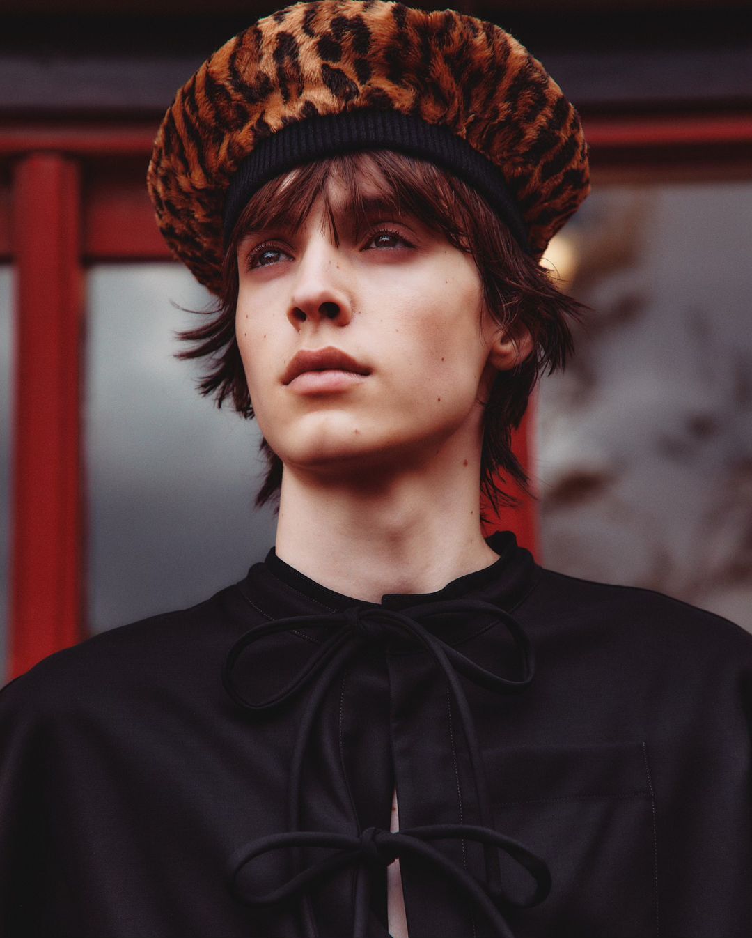 Portrait of a young adult with brown hair wearing a black top and leopard print beret, set against a blurred background of red and grey.