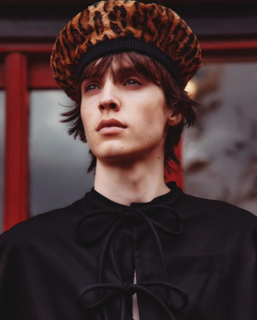 Portrait of a young adult with brown hair wearing a black top and leopard print beret, set against a blurred background of red and grey.