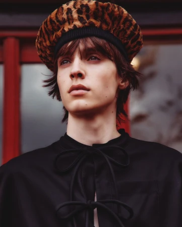 Portrait of a young adult with brown hair wearing a black top and leopard print beret, set against a blurred background of red and grey.