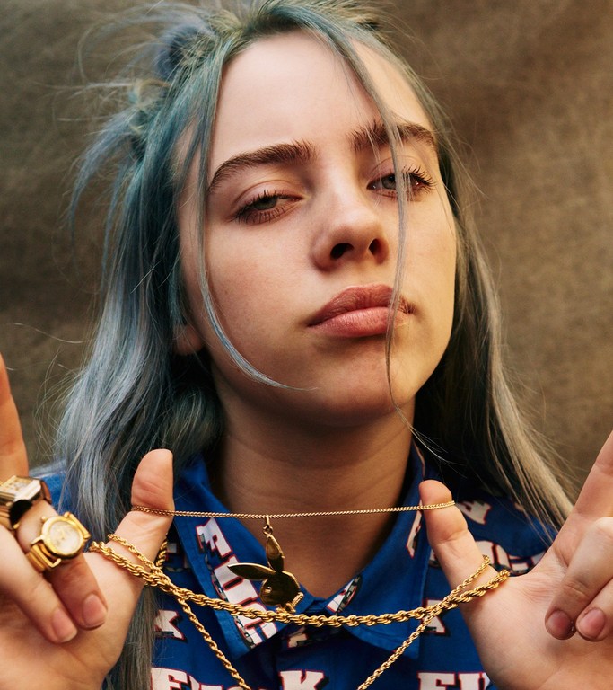 Close-up portrait of a young adult female with dyed blue hair, gold jewelry, and a patterned top against a neutral background.
